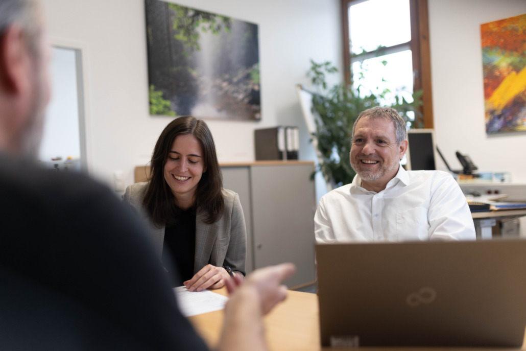 Drei Personen sitzen gemeinsam an einem Tisch in einem Büro und führen ein Gespräch. Zwei von ihnen lachen, eine Person hält ein Dokument in der Hand. Im Hintergrund sind Pflanzen und bunte Bilder zu sehen.