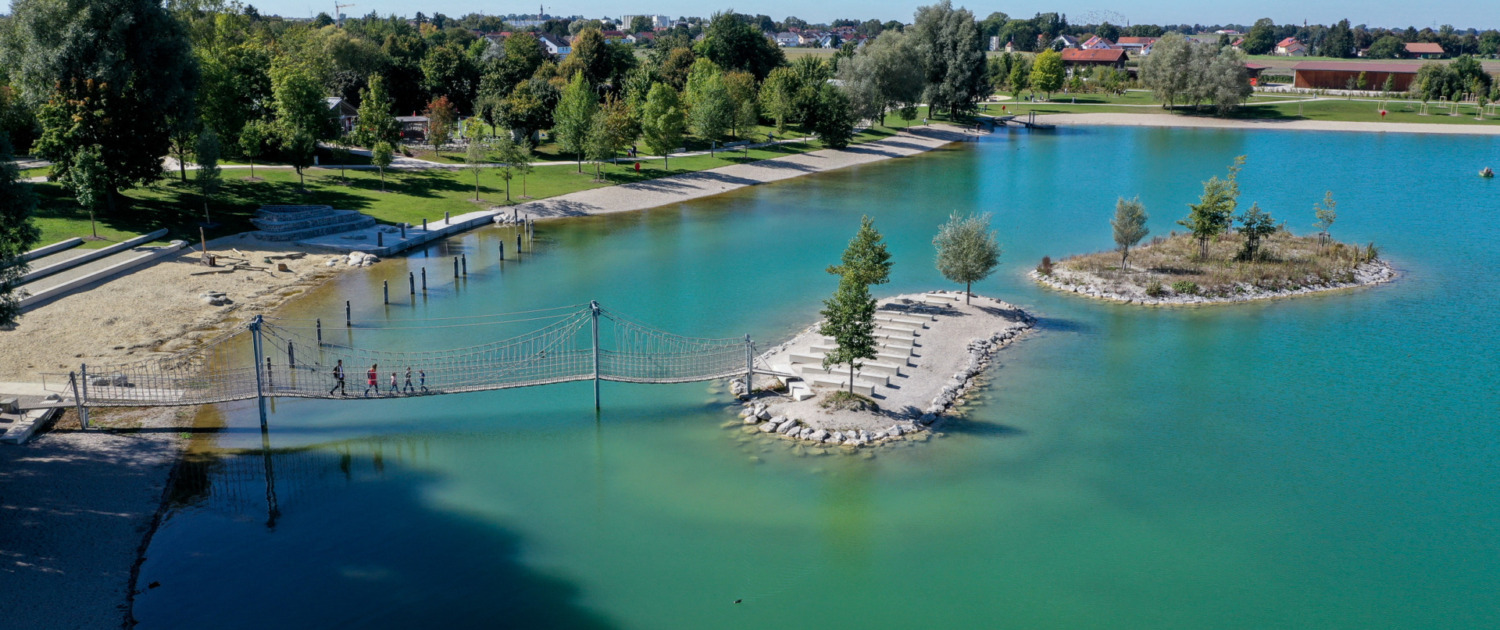 Luftaufnahme eines Badesees mit Hängebrücke, Inseln, Badebereich und umliegender Grünanlage.
