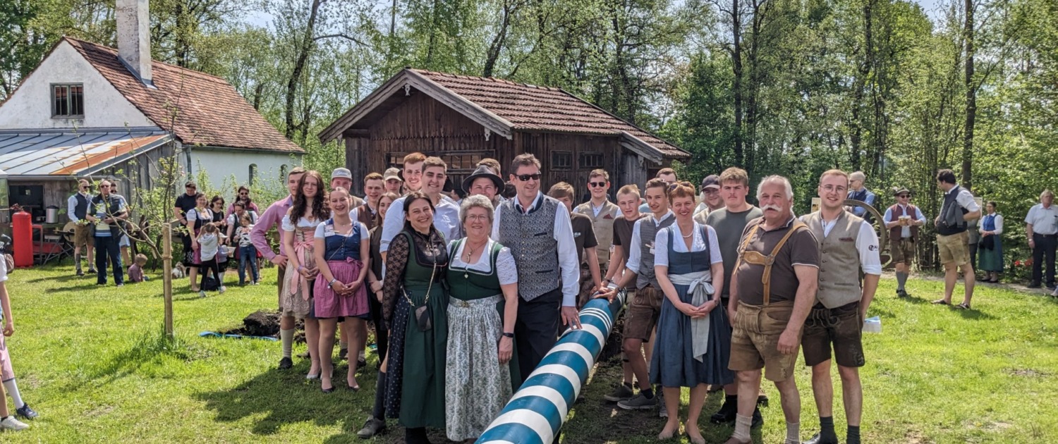 Menschen in Tracht versammeln sich beim traditionellen Maibaumaufstellen vor alten Bauernhäusern.