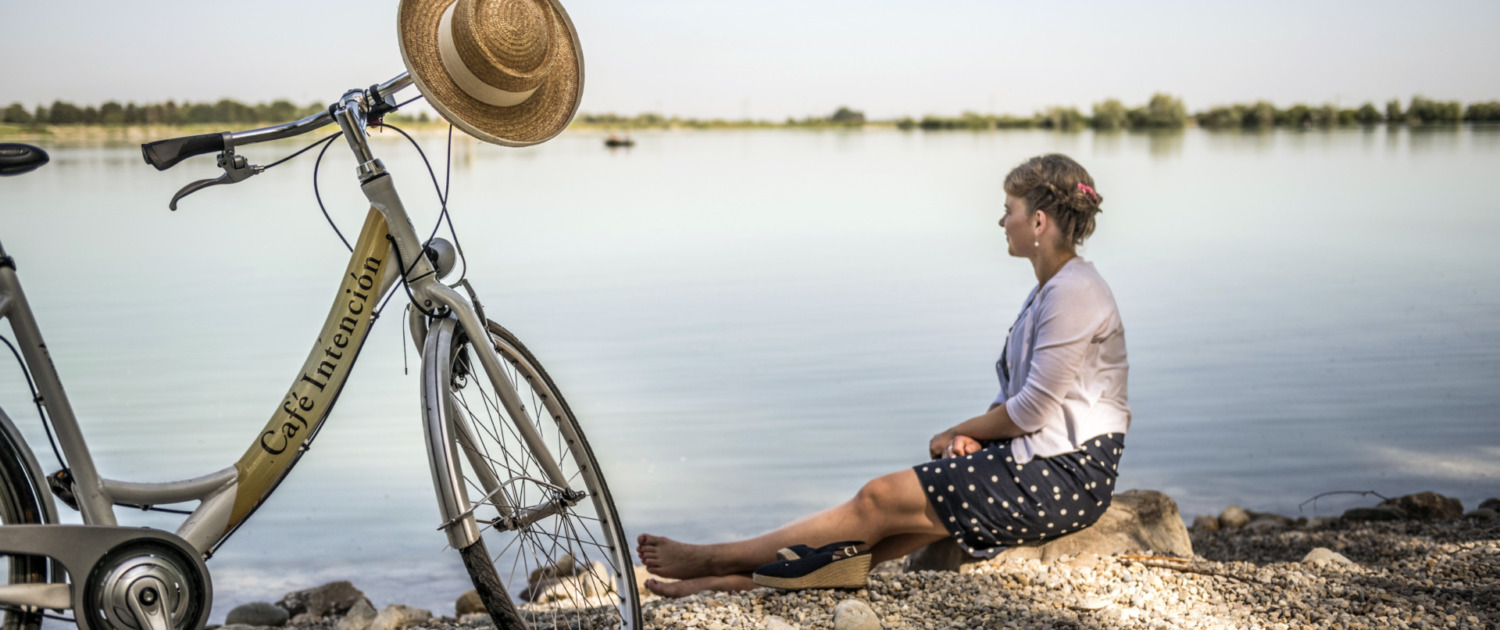 Eine Frau sitzt barfuß am Ufer eines Sees neben einem Fahrrad mit Strohhut am Lenker.