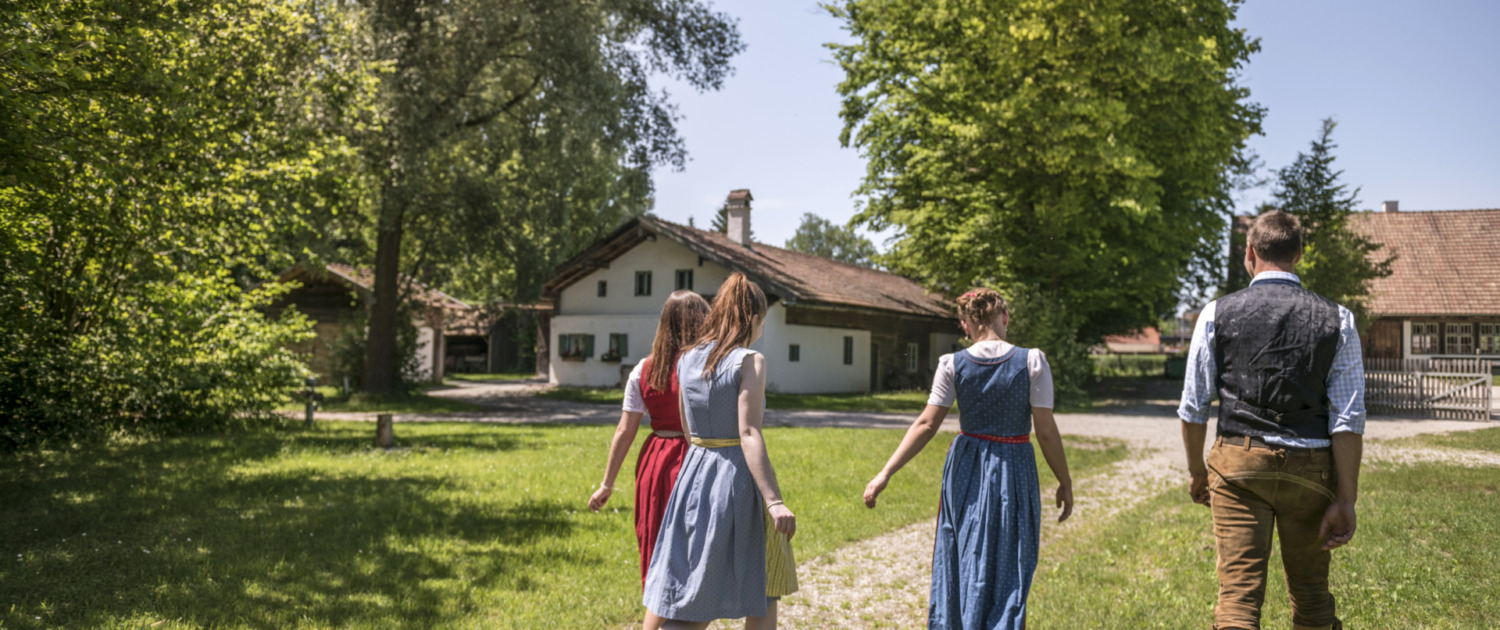 Vier Personen in Tracht gehen durch das Gelände eines historischen Bauernhausmuseums.