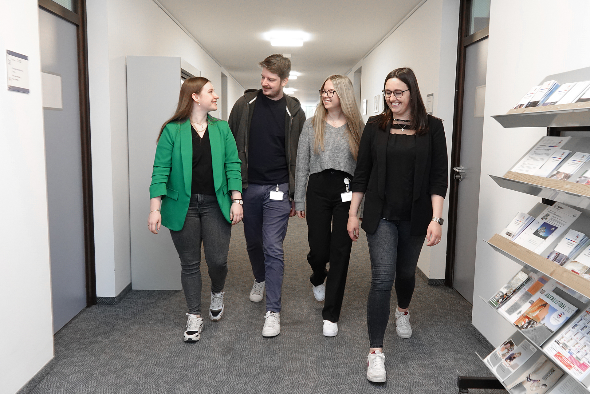 Vier junge Erwachsene gehen gemeinsam durch einen Büroflur und unterhalten sich freundlich. Rechts im Bild steht ein Prospektständer mit Informationsmaterial.