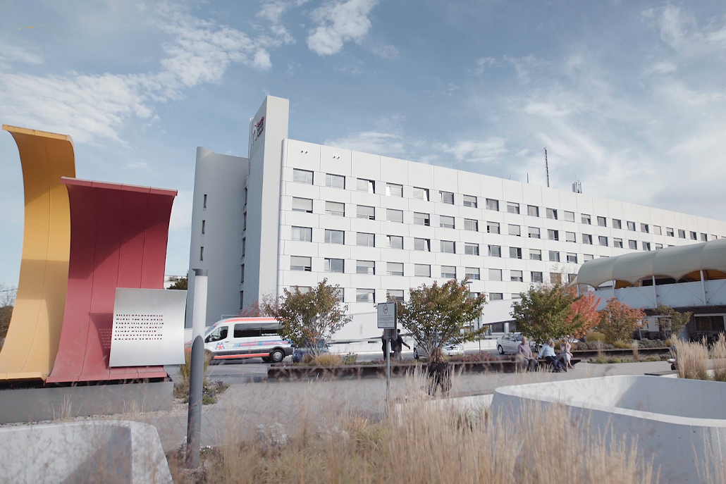 Außenansicht des mehrstöckigen Klinikums Landkreis Erding bei Tageslicht. Im Vordergrund sind Skulpturen, ein Gehweg, Bäume und ein Rettungswagen zu sehen.