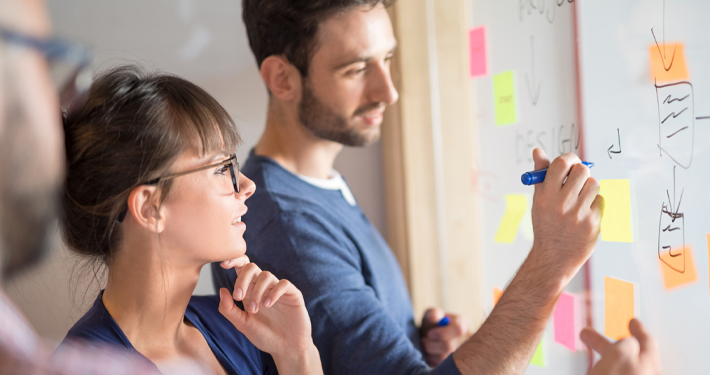 Zwei junge Menschen stehen vor einem Whiteboard mit bunten Klebezetteln und besprechen eine Idee.