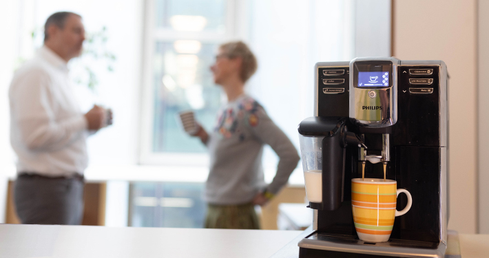 Im Vordergrund steht eine Kaffeemaschine mit einer Tasse darunter, im Hintergrund unterhalten sich zwei Personen mit Kaffeebechern.